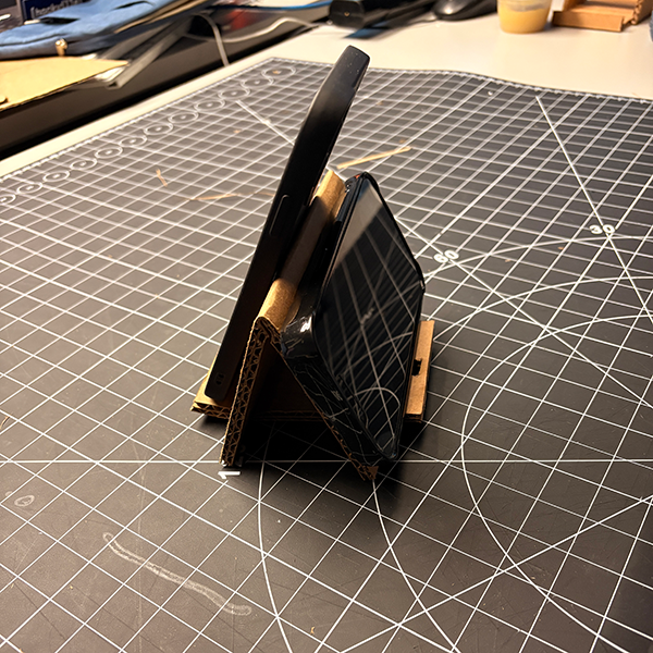 This is the final design of the phone stand that combines elements from both tutorials. It features a sturdy structure with a built-in phone charger hole, allowing users to charge their devices while using the stand. The dual-sided design accommodates two phones simultaneously, making it versatile for various use cases. This prototype exemplifies the principles of guerrilla prototyping by being quick to assemble, functional, and easily modifiable.